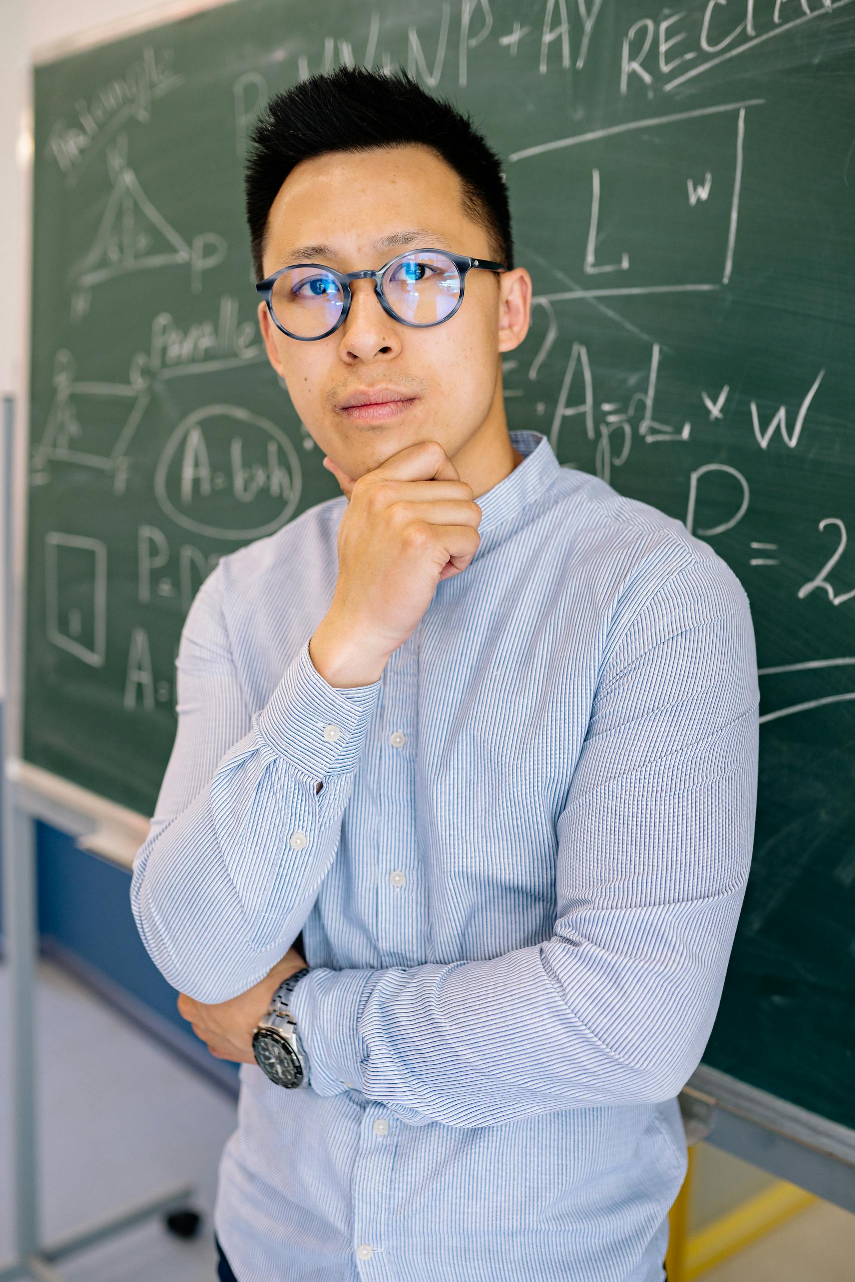 Male teacher standing thoughtfully in front of chalkboard filled with mathematical equations.