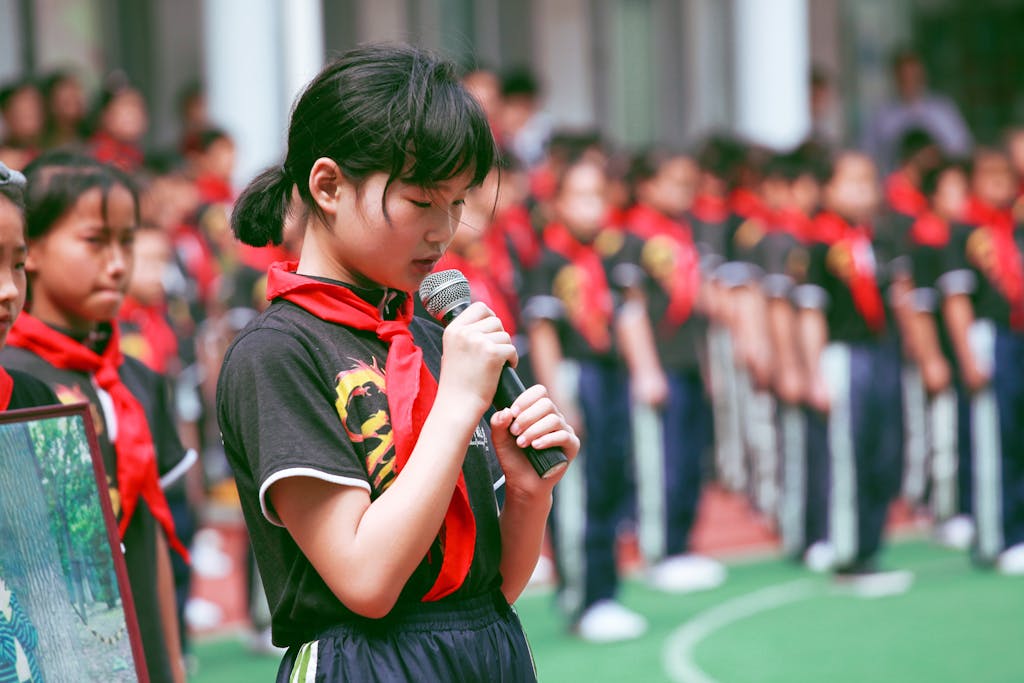 A young girl in uniform with a red scarf speaks into a microphone during an outdoor school event.