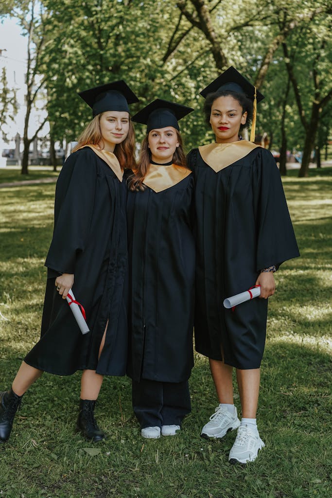Three diverse young women celebrating graduation outdoors in caps and gowns.